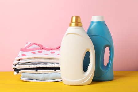 Bottles with fabric softeners and stack of baby clothes on yellow wooden table against pink backgroundの写真素材