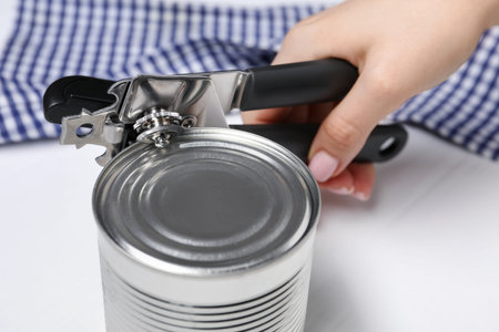 Woman opening tin with can opener at white table, closeup