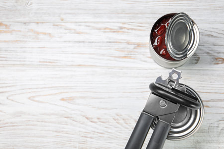 Can opener and tins on white wooden table, flat lay. Space for text