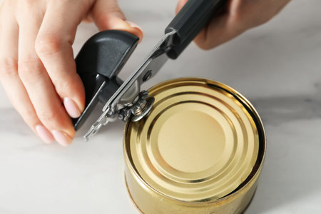 Woman opening tin with can opener at white table, closeup