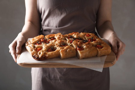 Woman holding cut delicious focaccia with tomatoes and spices on gray background, closeup