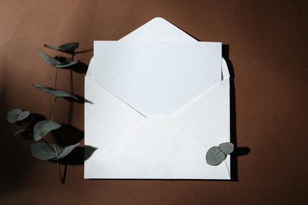 Blank greeting card in envelope and eucalyptus leaves on brown background, flat lay