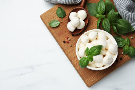 Tasty mozzarella cheese balls in bowl, spoon, basil and peppercorns on white marble table, flat lay. Space for textの写真素材