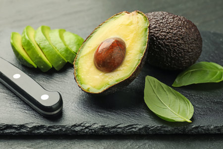 Tasty ripe green avocados, basil and knife on black table, closeup