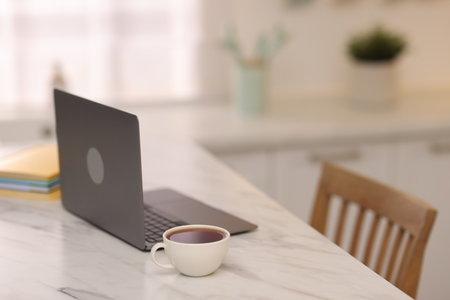 Laptop and cup of hot drink on white marble table in kitchen, selective focusの写真素材