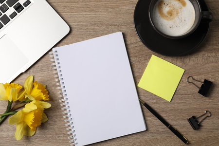 Notebook, other stationery, coffee, daffodils and laptop on wooden table, flat lay