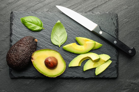 Tasty ripe green avocados, basil and knife on black table, flat lay
