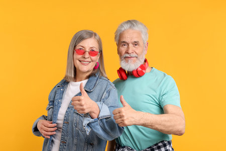 Senior couple in stylish outfits showing thumbs up on orange background