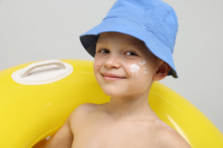 Little boy with sun protection cream on his skin and inflatable ring against light grey background