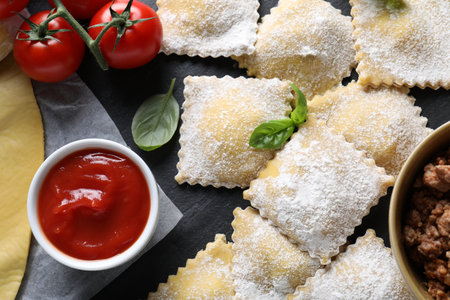 Uncooked ravioli, sauce, tomatoes and basil on black table, flat layの写真素材