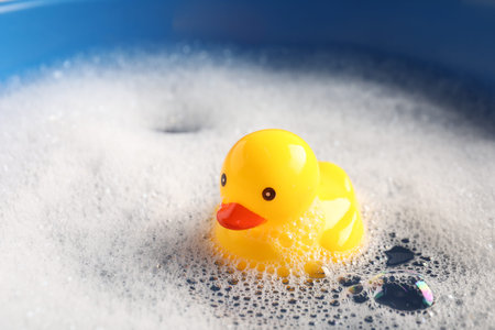 Cute rubber duck floating in water with bubbles against blue background, closeup