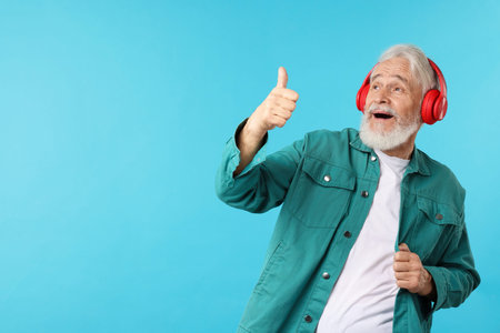 Elderly man in headphones showing thumbs up on light blue background, space for text