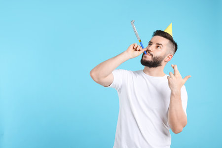 Happy Birthday. Handsome man with party hat and blower crossing fingers while making wish on light blue background, space for textの写真素材