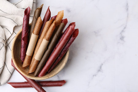 Tasty fruit leather rolls on white marble table, flat lay. Space for text