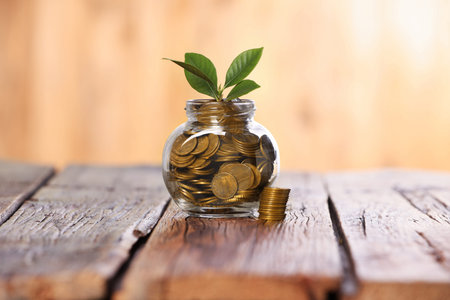 Investment concept. Glass jar with coins and sprout on wooden table against blurred orange backgroundの写真素材