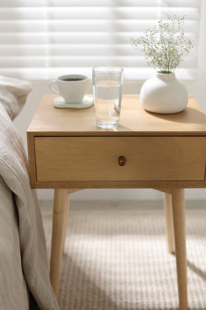 Bedside table with glass of water, flowers in vase and coffee near bed indoors