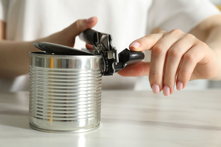 Woman opening tin with can opener at white table, closeup