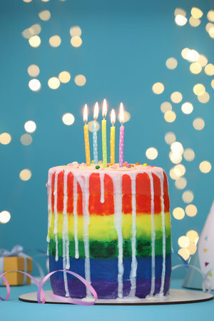 Delicious rainbow birthday cake with burning candles and streamers on light blue table against blurred lights, closeupの写真素材