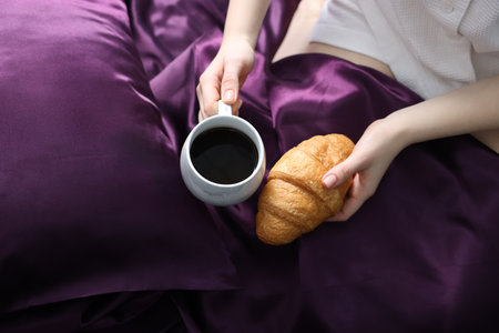 Woman with coffee and croissant on bed with purple silk linens, top viewの写真素材