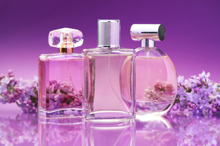Bottles with perfumes and lilac flowers on mirror surface against color gradient background, closeup