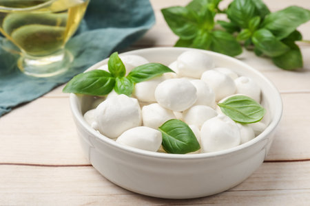 Tasty mozzarella cheese balls in bowl and basil on light wooden table, closeupの写真素材
