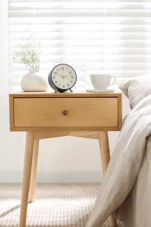 Bedside table with alarm clock, flowers in vase, cup and glass of water near bed indoorsの写真素材