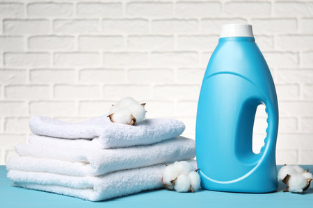 Bottle of fabric softener, towels and cotton flowers on light blue table against white brick wallの写真素材