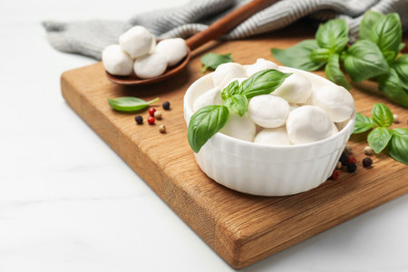 Tasty mozzarella cheese balls in bowl, spoon, basil and peppercorns on white marble table, closeupの写真素材
