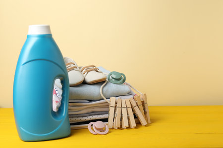Bottle with fabric softener, baby clothes, pacifiers and clothespins on yellow wooden table against beige background. Space for textの写真素材
