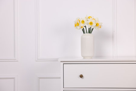 Beautiful daffodils in vase on dresser near white wall at home, space for textの写真素材