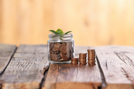 Investment concept. Glass jar with coins and sprout on wooden table against blurred orange...