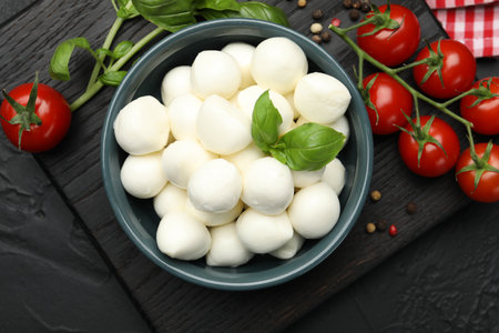 Tasty mozzarella cheese balls in bowl, basil, peppercorns and tomatoes on black table, top viewの写真素材