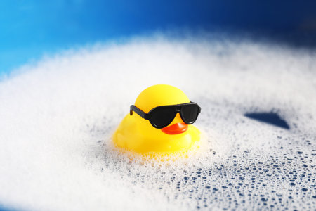 Cute rubber duck floating in water with bubbles against blue background, closeup