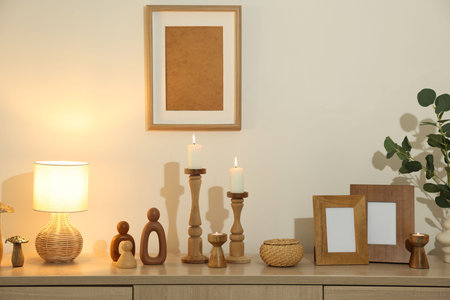 Stylish candlesticks with burning candles and other decor on cabinet near beige wall indoors