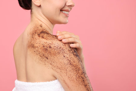Smiling woman applying body scrub onto her shoulder against pink background, closeup. Space for text