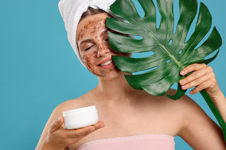 Smiling woman with body scrub on her face, jar and monstera leaf against light blue backgroundの写真素材