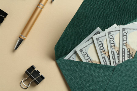 Money bonus. Envelope with dollar banknotes, pen and binder clip on beige background, flat lay