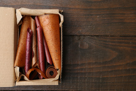 Delicious fruit leather rolls in cardboard box on wooden table, top view. Space for textの写真素材