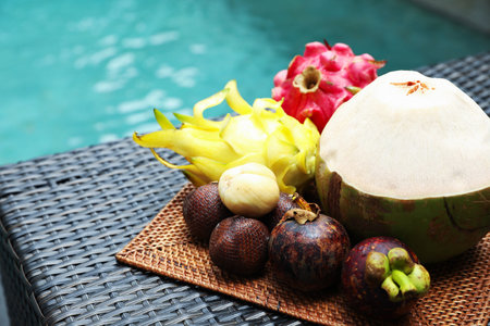 Different tropical fruits on wicker table near swimming pool, closeup. Space for textの写真素材