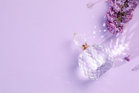 Bottle of perfume and beautiful lilac flowers on light violet background, flat lay. Space for text
