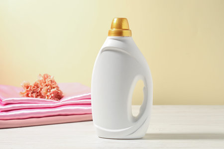Bottle of fabric softener, clean clothes and carnation flowers on white wooden table against light...