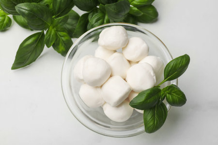 Tasty mozzarella cheese balls in bowl and basil on white marble table, flat lay