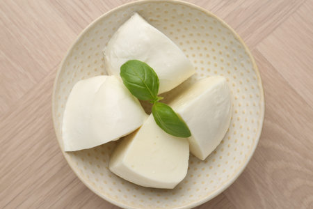 Pieces of delicious mozzarella cheese and basil in bowl on wooden table, top view