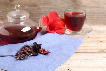 Delicious hibiscus tea in glass cup, teapot, dry roselle sepals and spoon on wooden table, closeup. Space for textの写真素材