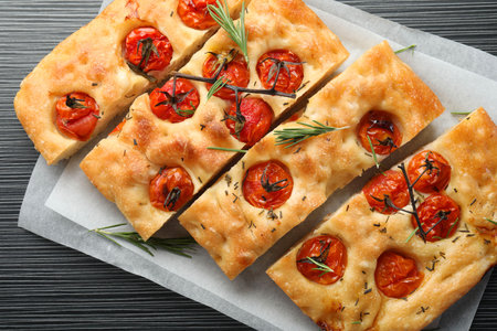 Cut delicious focaccia with tomatoes and spices on dark wooden table, top view