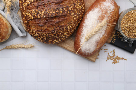 Fresh bread loaves and spikes on white tiled table, flat lay. Space for text