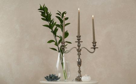 Elegant candlestick with burning candles, branches in vase and other decor on white table near beige...
