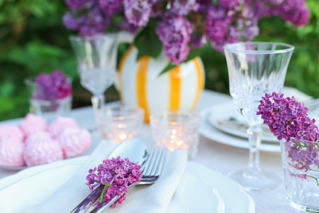 Beautiful lilac flowers, zephyrs, plates, cutlery, glasses and burning candles on table in garden,...