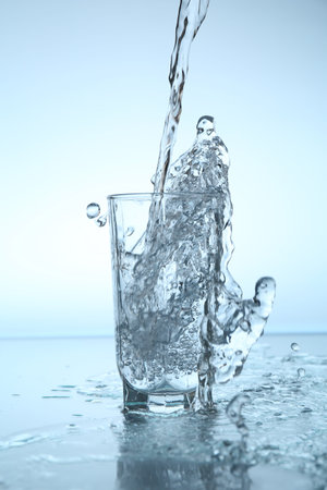 Pouring water into glass at mirror table against light blue gradient background, closeup