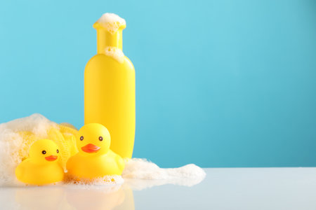 Cute rubber ducks, foam, sponge and bottle of shampoo on table against light blue background, space for textの写真素材